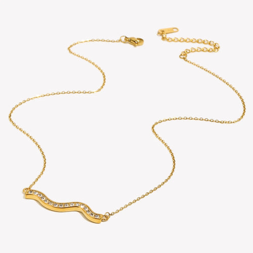 Gold pavé wave bar necklace on light grey-white background, overhead angled view showing full chain length with lobster clasp and extender at top, rhinestone wave pendant at bottom, fine O-chain links visible
