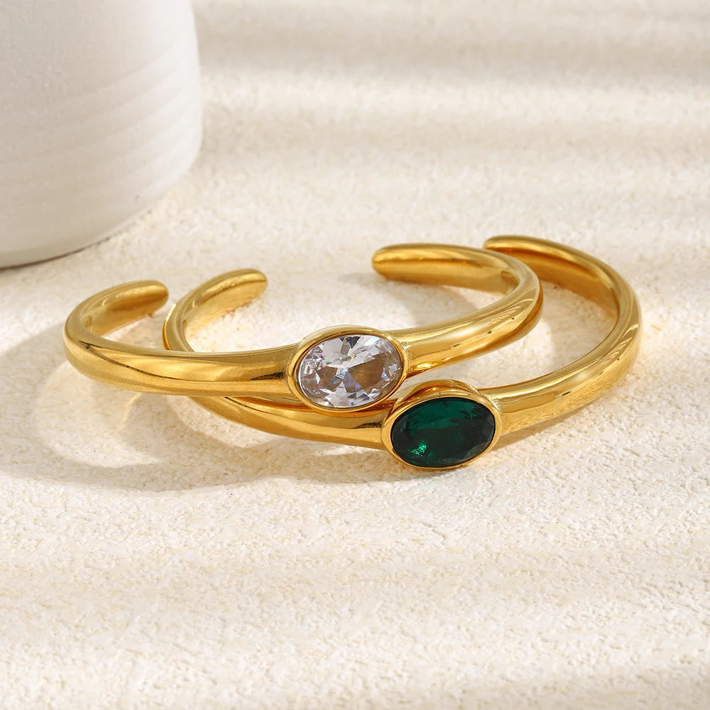 Gold oval stone open cuff bracelets in clear and emerald green stacked on cream fabric surface, bezel-set oval stones and smooth polished band in warm directional light, white ceramic object in background
