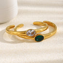 Gold oval stone open cuff bracelets in clear and emerald green stacked on cream fabric surface, bezel-set oval stones and smooth polished band in warm directional light, white ceramic object in background
