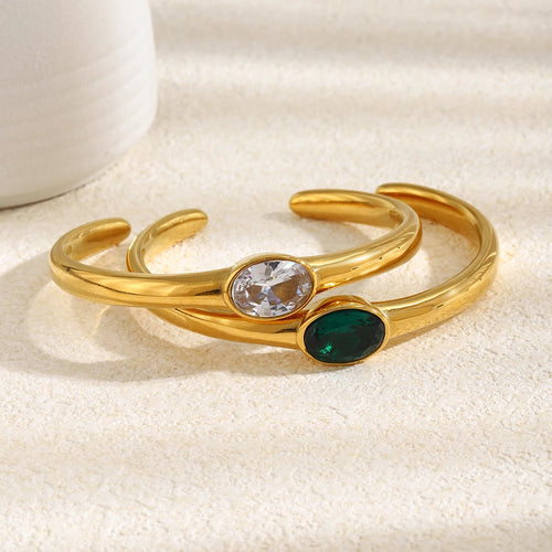Gold oval stone open cuff bracelets in clear and emerald green stacked on cream fabric surface, bezel-set oval stones and smooth polished band in warm directional light, white ceramic object in background
