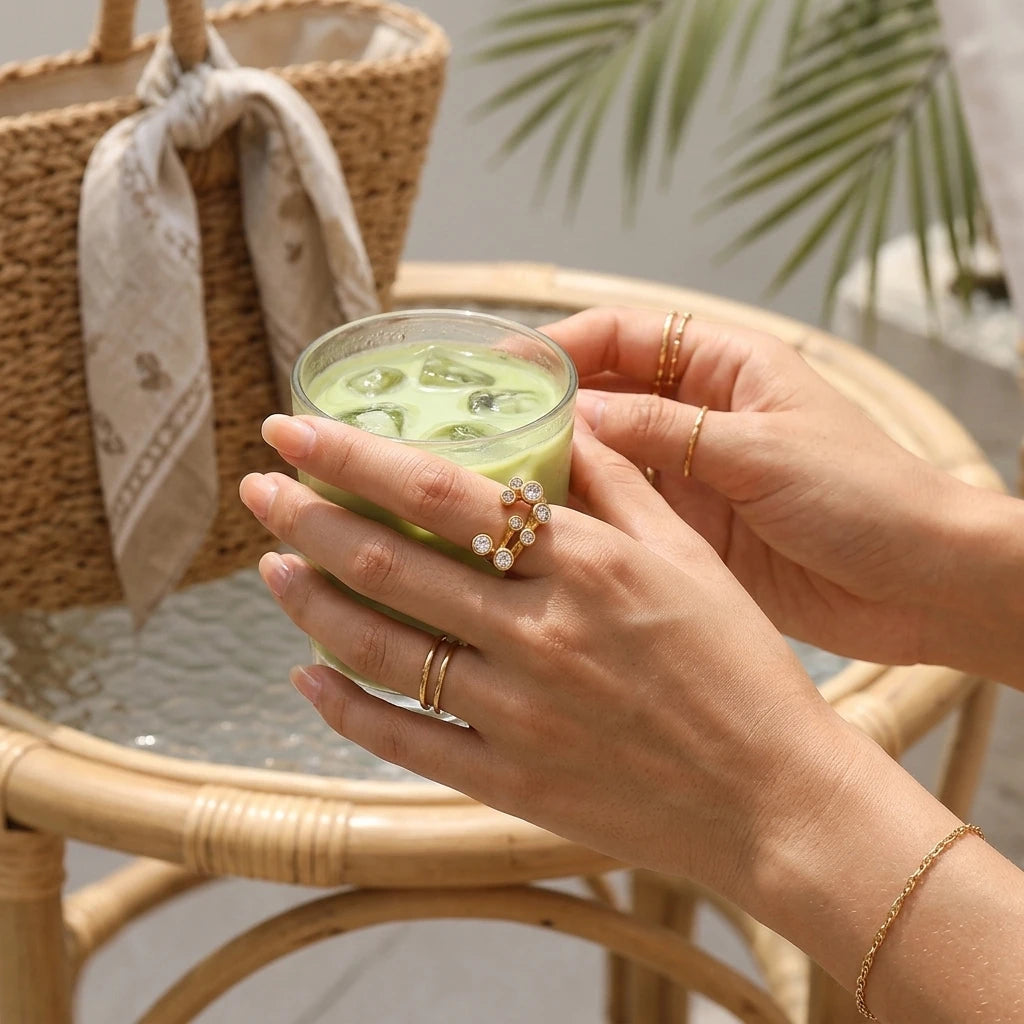 Woman holding iced matcha glass wearing gold clear stone scatter open band ring on middle finger, rattan chair and palm leaf in background, gold chain bracelet visible
