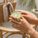 Woman holding iced matcha glass wearing gold clear stone scatter open band ring on middle finger, rattan chair and palm leaf in background, gold chain bracelet visible
