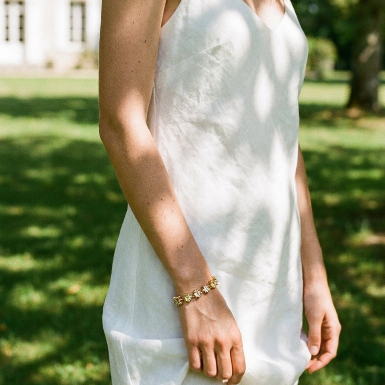 Woman wearing gold multicolor square stone tennis bracelet on wrist, arm hanging naturally beside white silk slip dress, dappled sunlight on lawn, blurred green garden and manor house background
