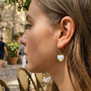Woman wearing gold iridescent heart drop hoop earrings at outdoor European cafe, side profile with straight brown hair, domed iridescent heart charm visible below ear, stone street and wicker chair in background
