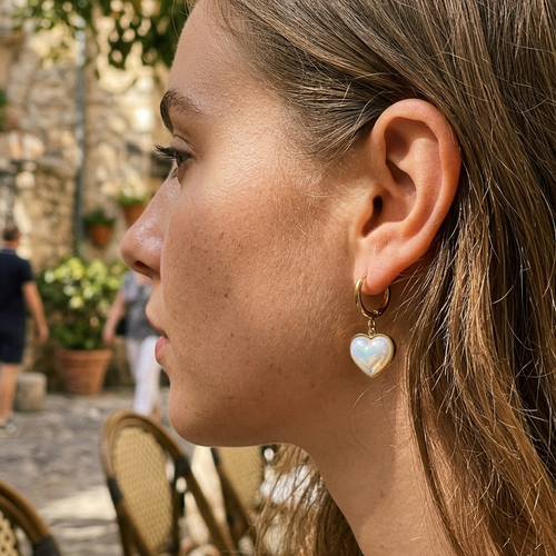 Woman wearing gold iridescent heart drop hoop earrings at outdoor European cafe, side profile with straight brown hair, domed iridescent heart charm visible below ear, stone street and wicker chair in background
