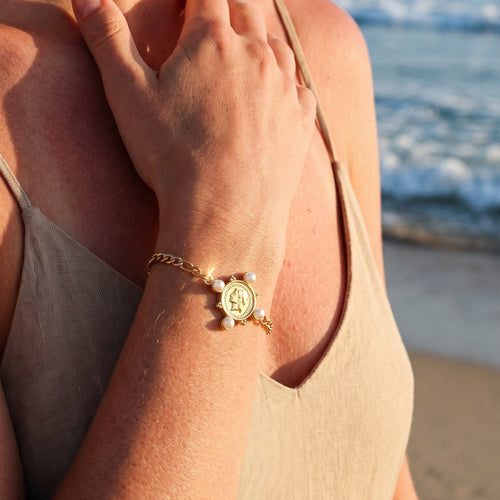Woman wearing gold pearl coin charm Figaro chain bracelet at sunset beach, hands crossed over chest, beige linen tank top, coin charm and pearl accents catching warm golden light, blurred ocean waves in background
